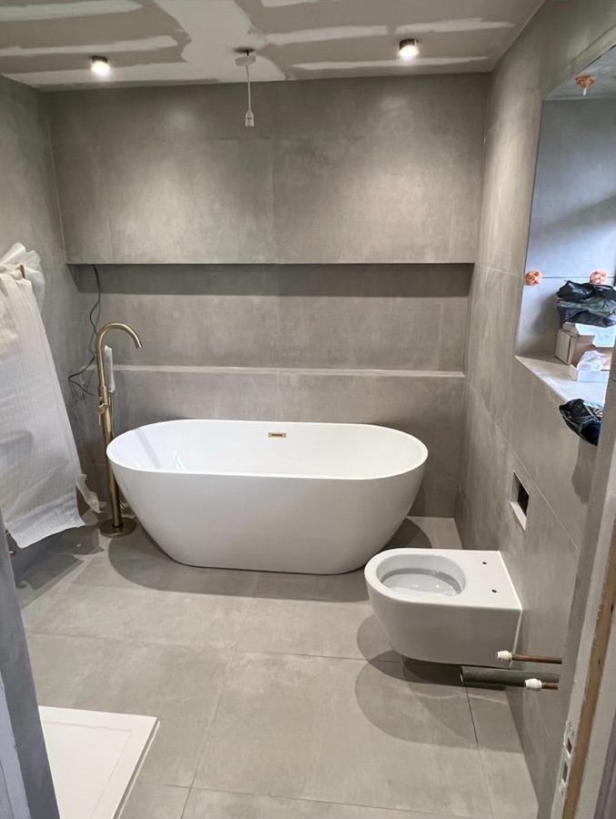Concrete effect tiled bathroom with freestanding bath and brass tap during installation