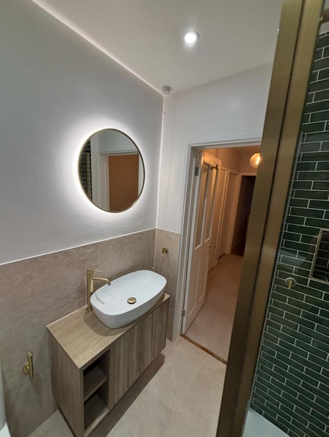 Oak vanity with brass tap and backlit round mirror beside a forest green metro shower